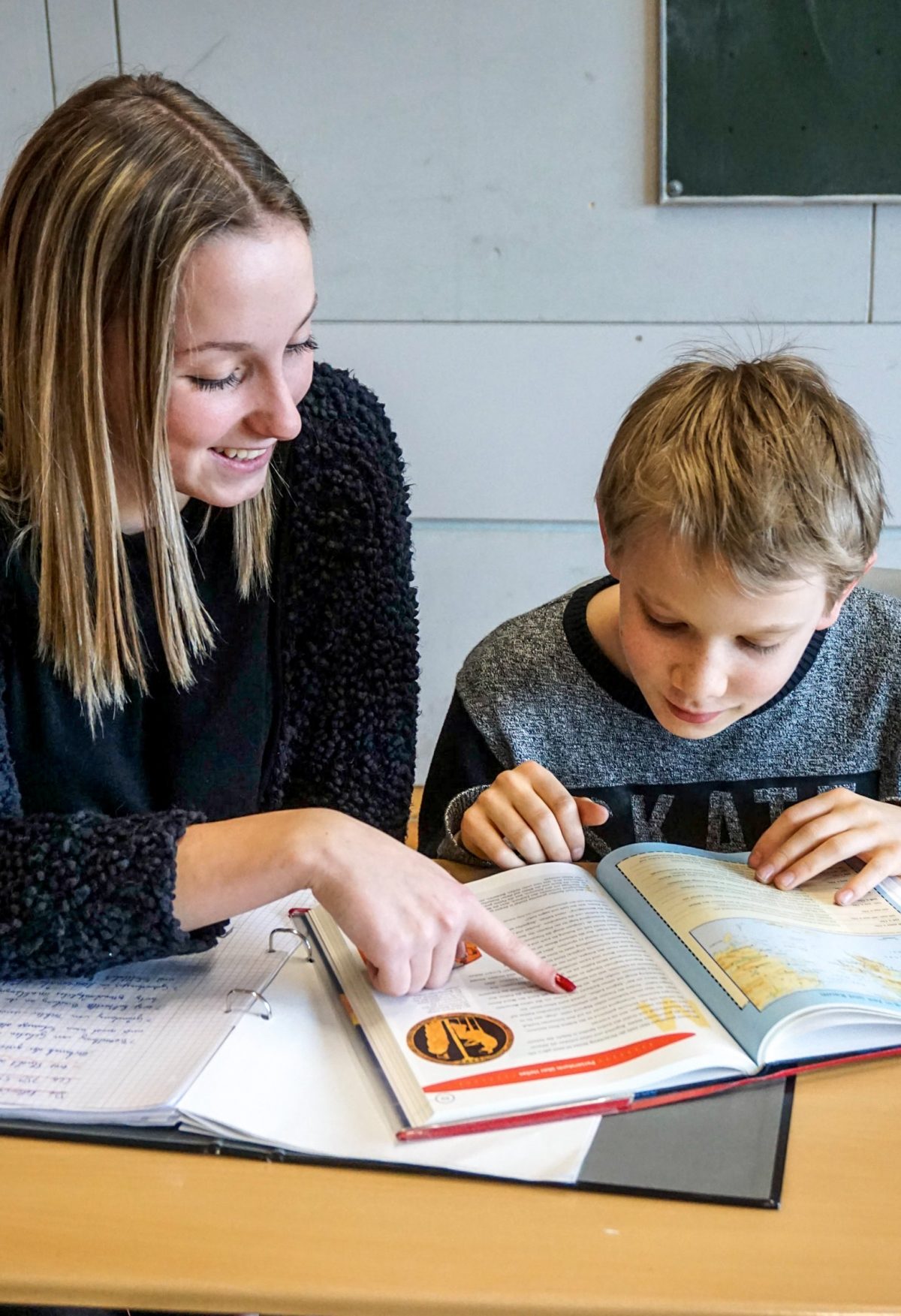 Eine ältere Schülerin sitzt mit einem jüngeren Schüler an einem Tisch und zeigt ihm etwas in einem Buch